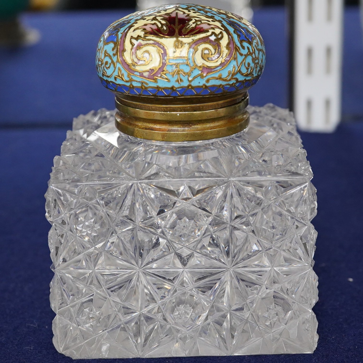 A large square glass inkwell, heavily cut with star cut base and octagonally cut top with enamelled brass hinged cover, 8cm wide. Condition - fair, some chipping